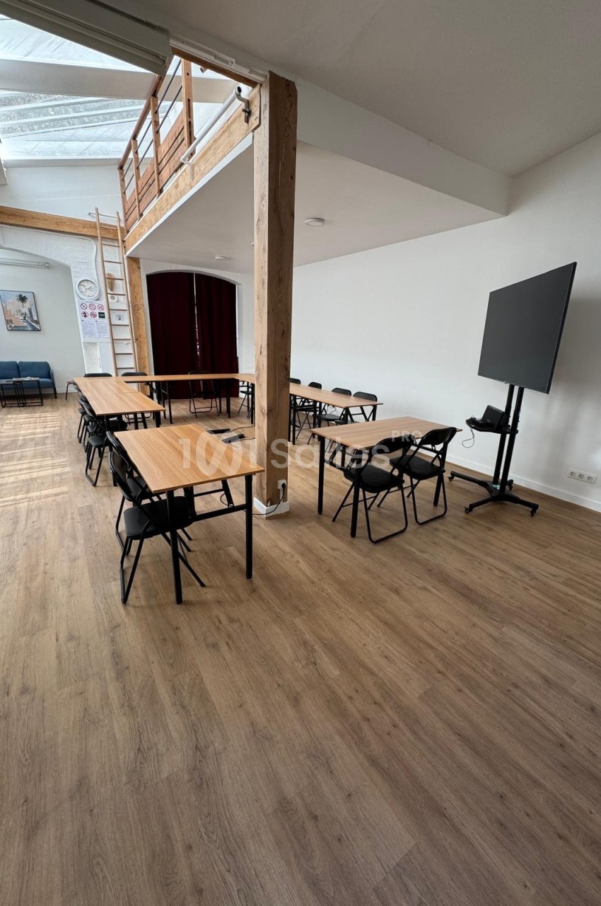 Salle lumineuse avec parquet, tables, chaises, écran sur pied et mezzanine en bois visible en arrière-plan.