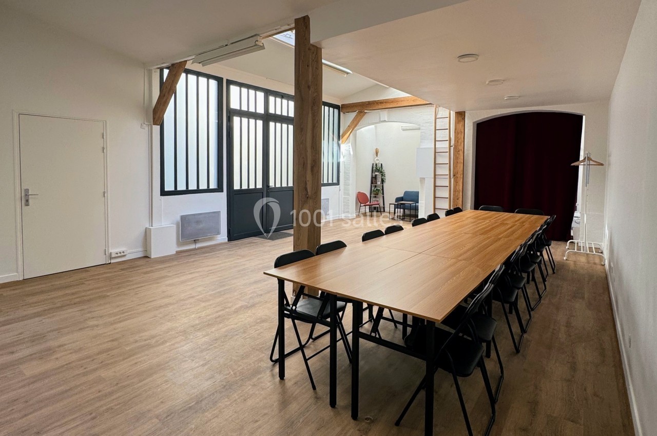 Salle lumineuse avec parquet, grande table en bois entourée de chaises noires, poutres apparentes et verrière.