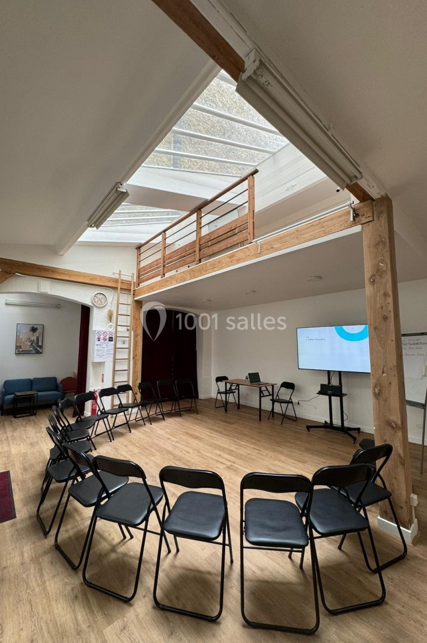 Salle lumineuse avec chaises disposées en cercle, écran de présentation, tableau blanc et mezzanine en bois.