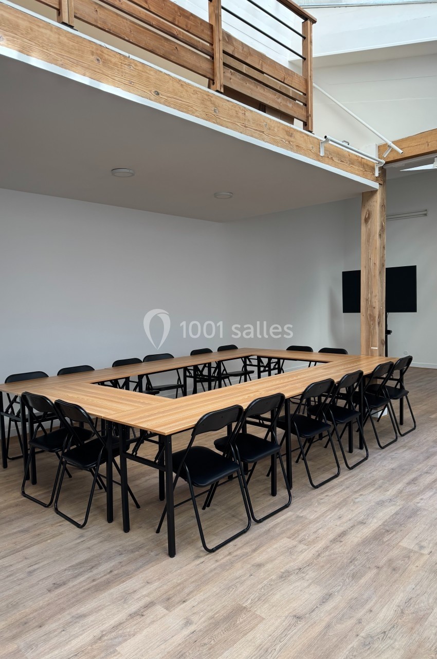 Salle de réunion avec une grande table en U entourée de chaises noires, dans un espace lumineux avec parquet et mezzanine en…