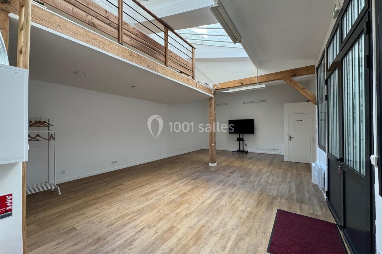 Salle lumineuse avec parquet, mezzanine en bois, grande porte vitrée et écran noir au fond.