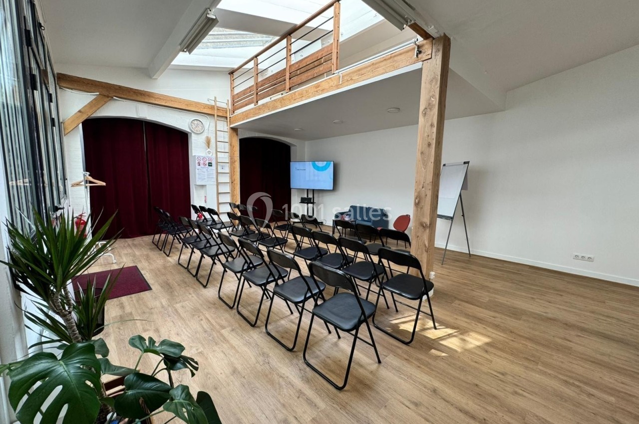 Salle lumineuse avec parquet, chaises alignées face à un écran, mezzanine en bois et grandes baies vitrées.