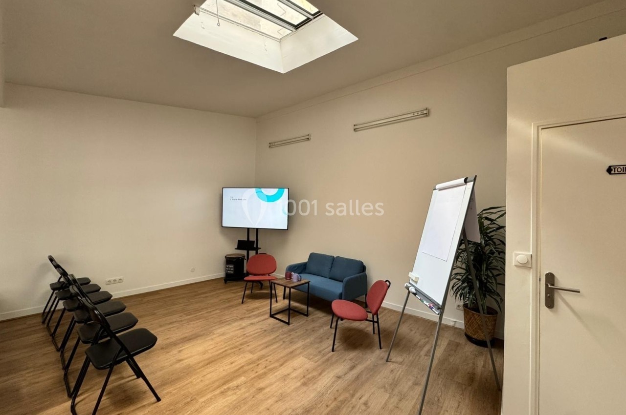 Salle de réunion lumineuse avec chaises pliantes, canapé, écran, tableau blanc et plante en pot.