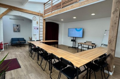 Salle lumineuse avec parquet, grande table entourée de chaises, écran, paperboard et mezzanine en bois.