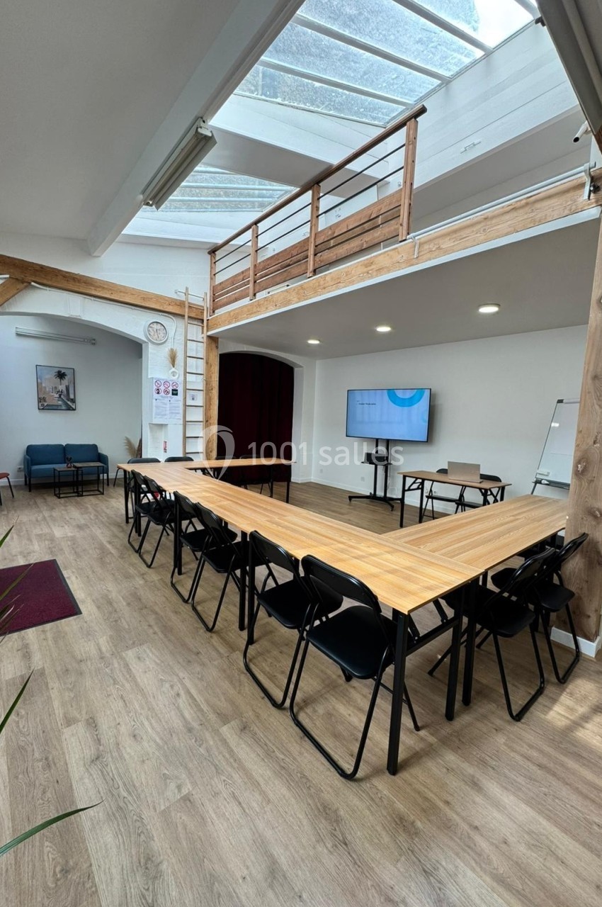 Salle de réunion lumineuse avec grande table en U, chaises noires, écran mural et mezzanine en bois.