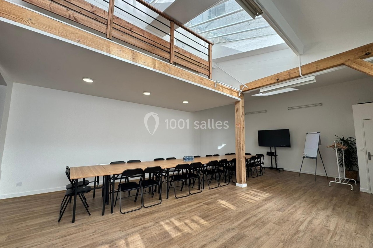 Salle lumineuse avec parquet, grande table entourée de chaises, écran, paperboard et mezzanine en bois.