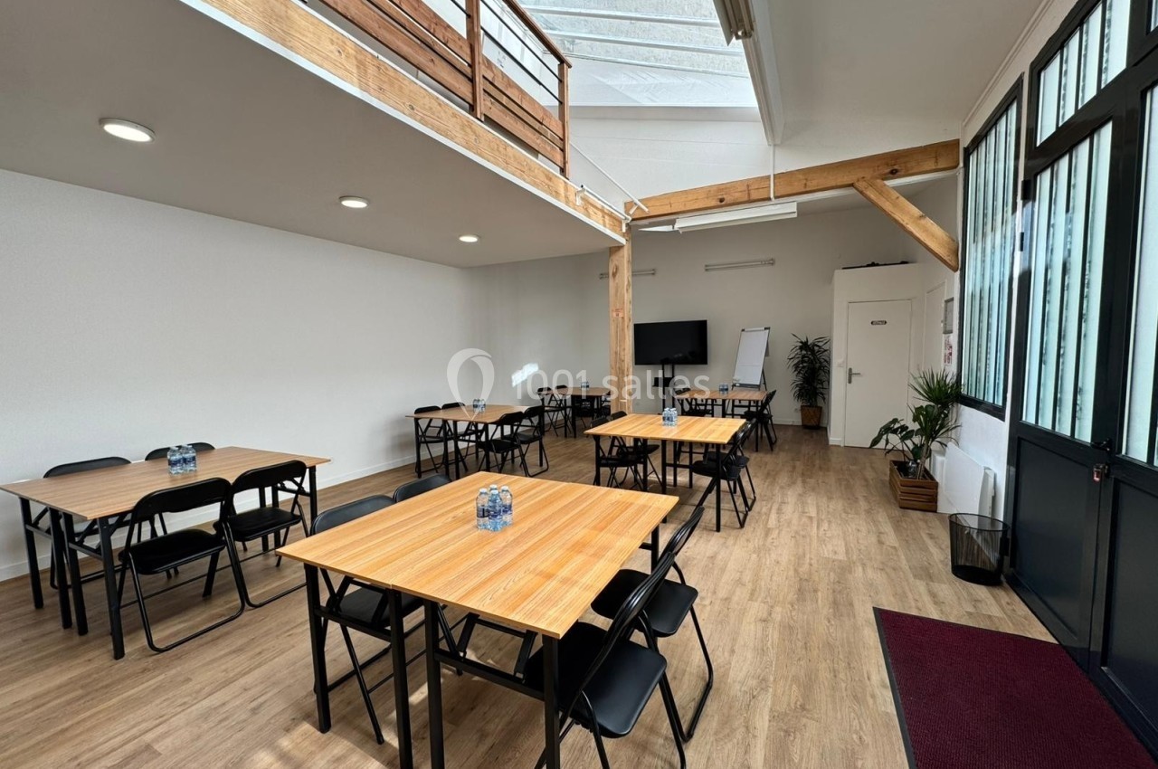 Salle lumineuse avec tables en bois, chaises noires, tableau blanc, plantes et mezzanine en bois.