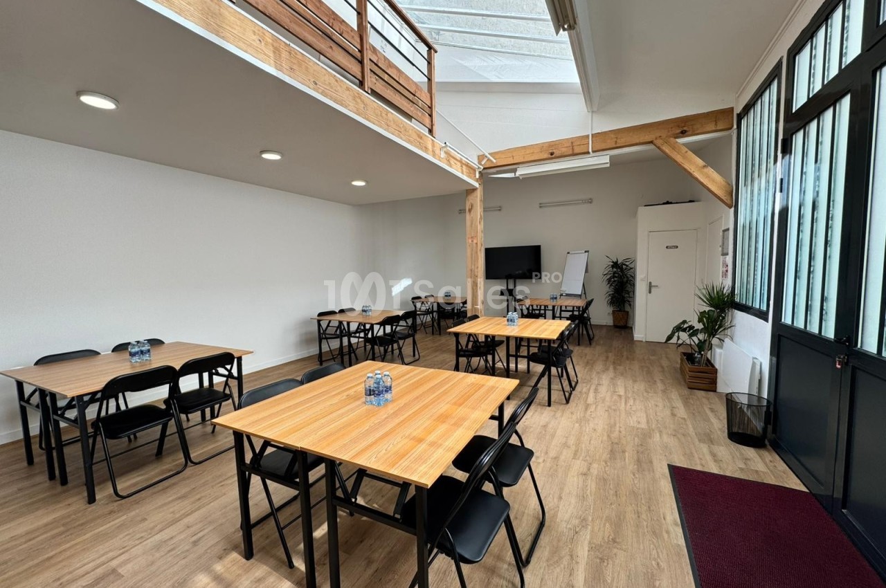 Salle lumineuse avec tables en bois, chaises noires, tableau blanc, plantes et mezzanine en bois.