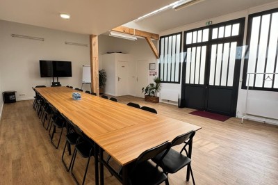 Salle lumineuse avec parquet, grande table entourée de chaises, écran, paperboard et mezzanine en bois.