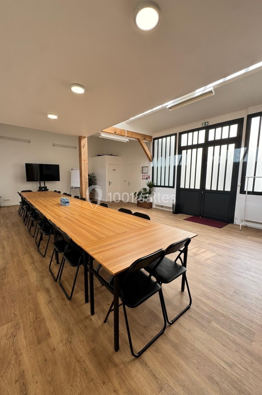 Salle de réunion lumineuse avec une grande table en bois, des chaises noires, un écran et des portes vitrées.