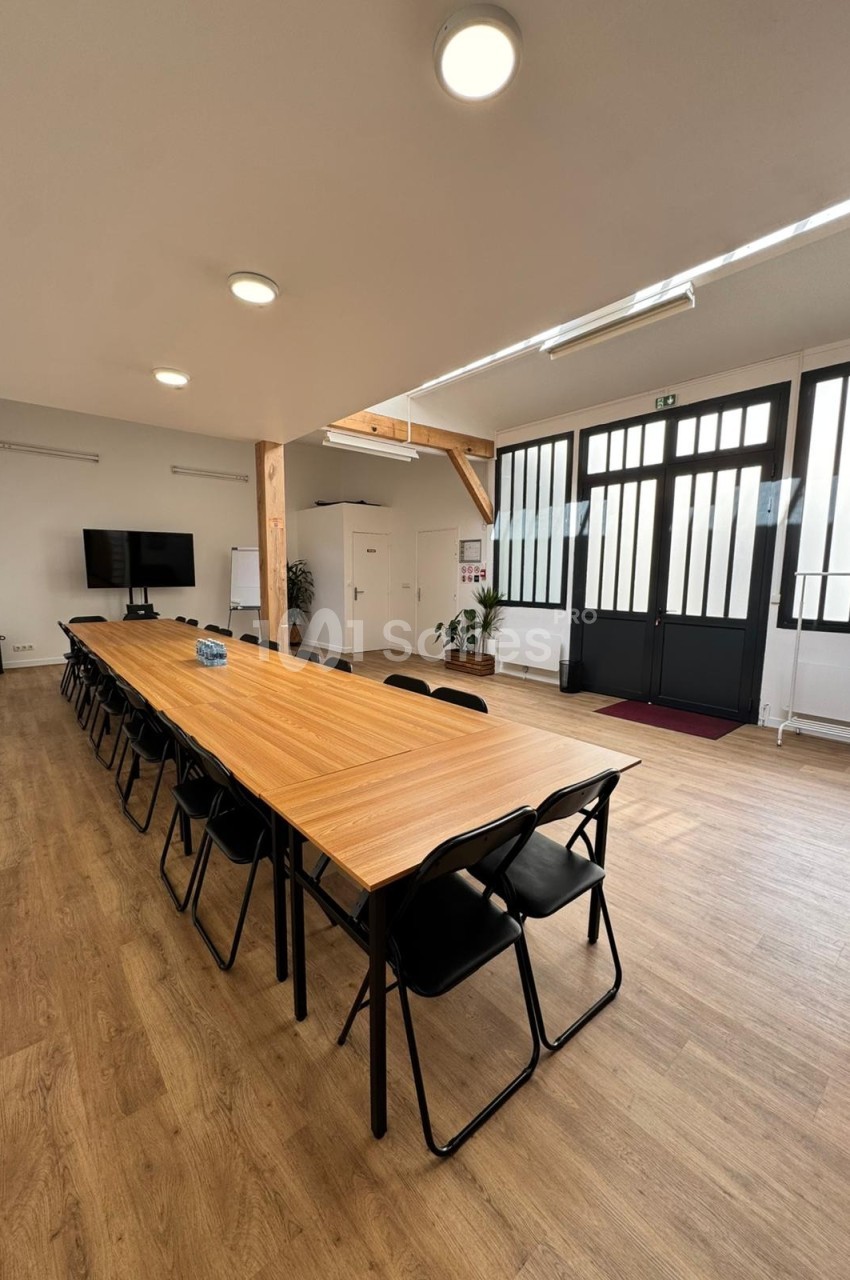 Salle de réunion lumineuse avec une grande table en bois, des chaises noires, un écran et des portes vitrées.
