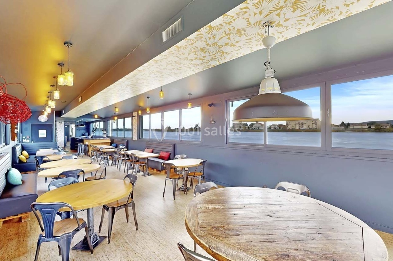 Salle lumineuse avec grandes fenêtres, tables en bois rondes, chaises métalliques et décorations modernes.