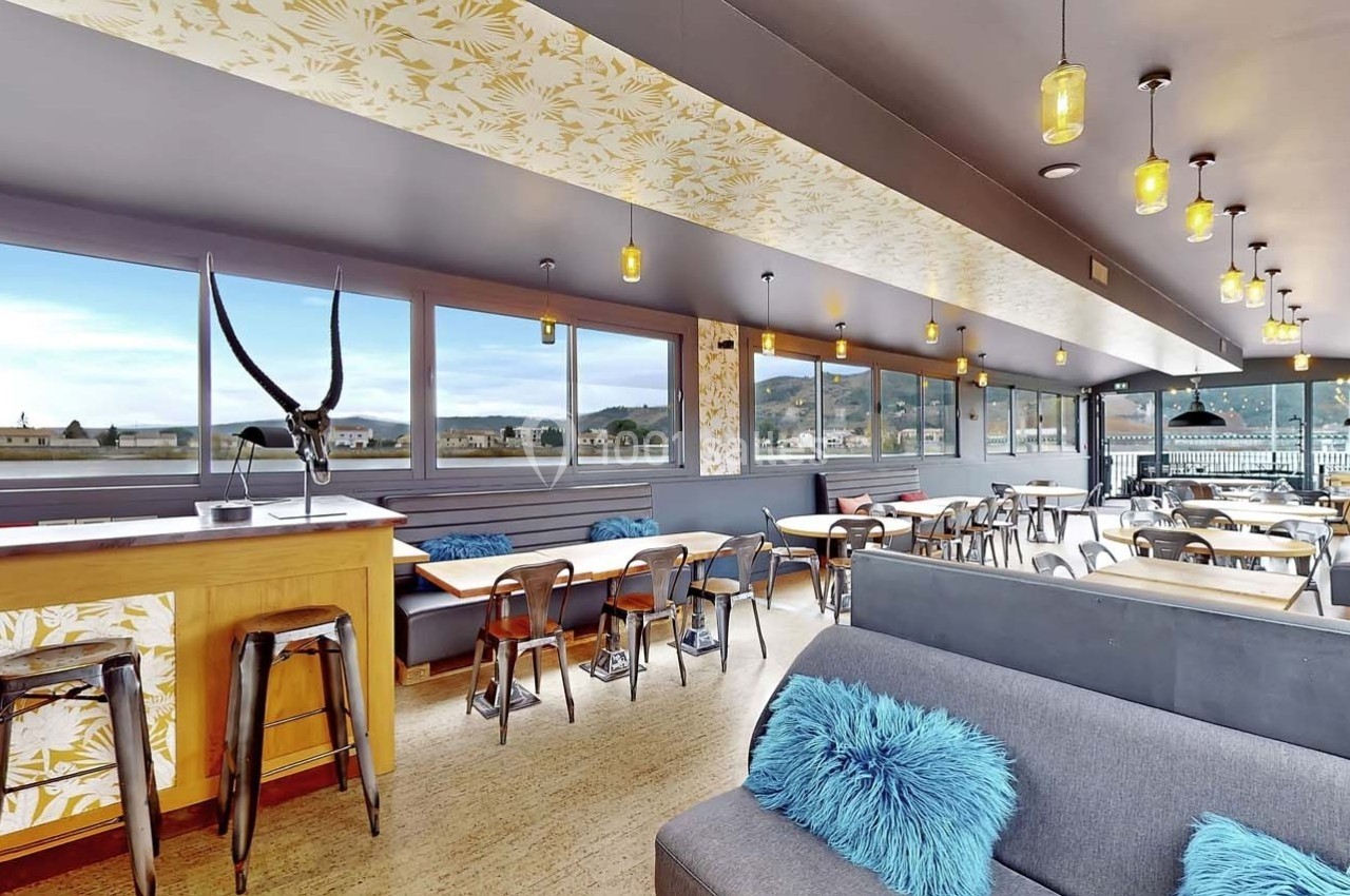 Salle lumineuse avec de grandes baies vitrées, mobilier en bois, banquettes grises et décorations modernes.