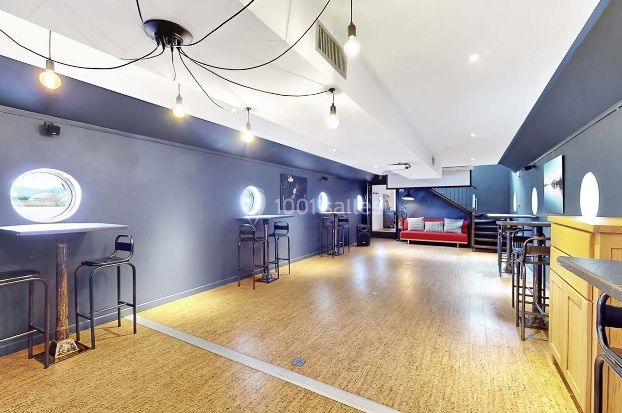 Salle lumineuse avec parquet, murs bleus, tables hautes, tabourets, hublots et éclairage suspendu.