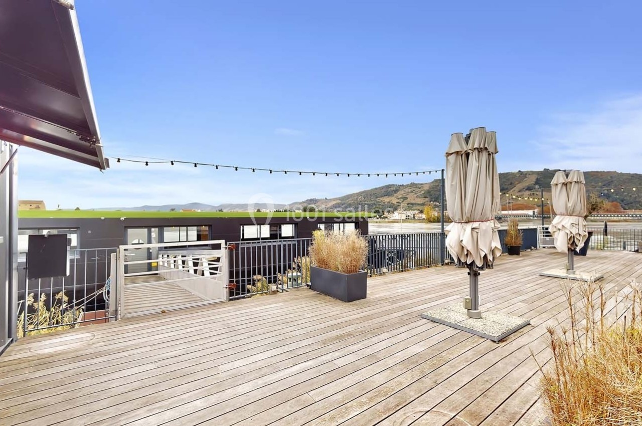 Terrasse en bois avec parasols fermés, plantes en pots et vue sur des collines sous un ciel dégagé.