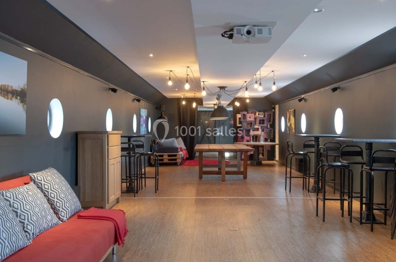 Salle aménagée avec des tables, chaises hautes, canapés et éclairage suspendu, dans un espace aux murs sombres avec hublots.