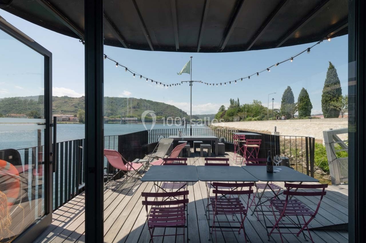 Terrasse en bois avec tables et chaises colorées, vue sur une rivière et collines sous un ciel dégagé.