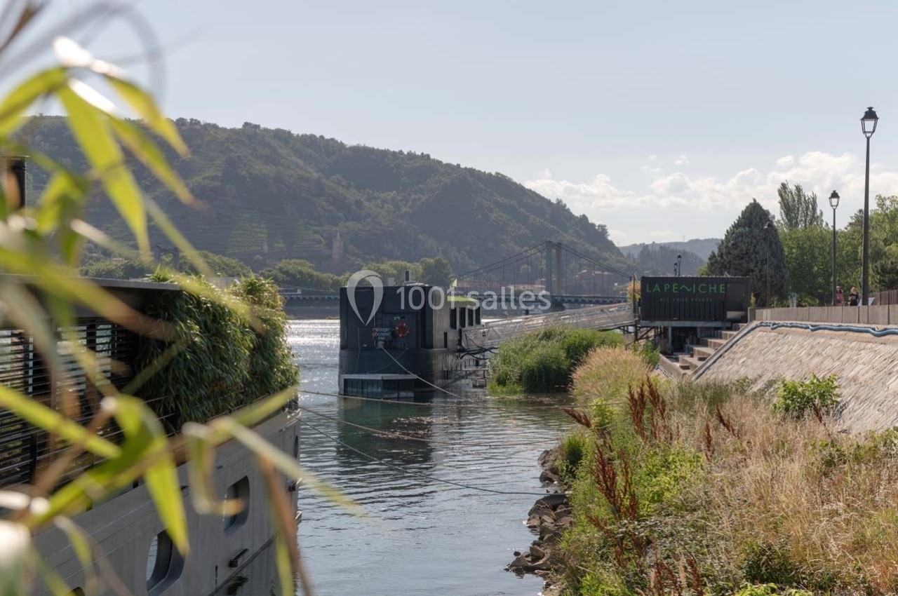 Vue d'une rivière bordée de végétation, avec une péniche amarrée et un pont visible à l'arrière-plan.