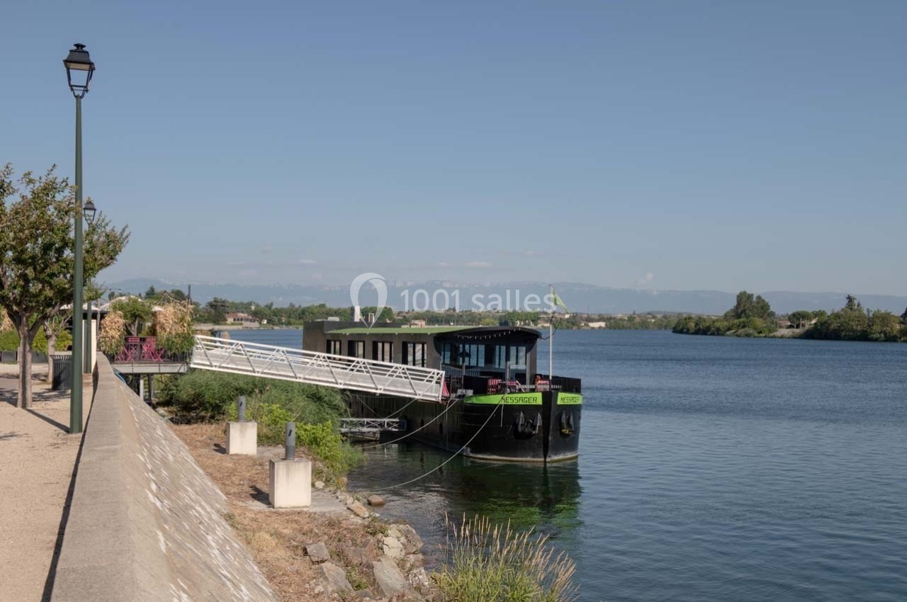 Péniche amarrée le long d'une berge arborée, avec un paysage de rivière et des montagnes en arrière-plan.