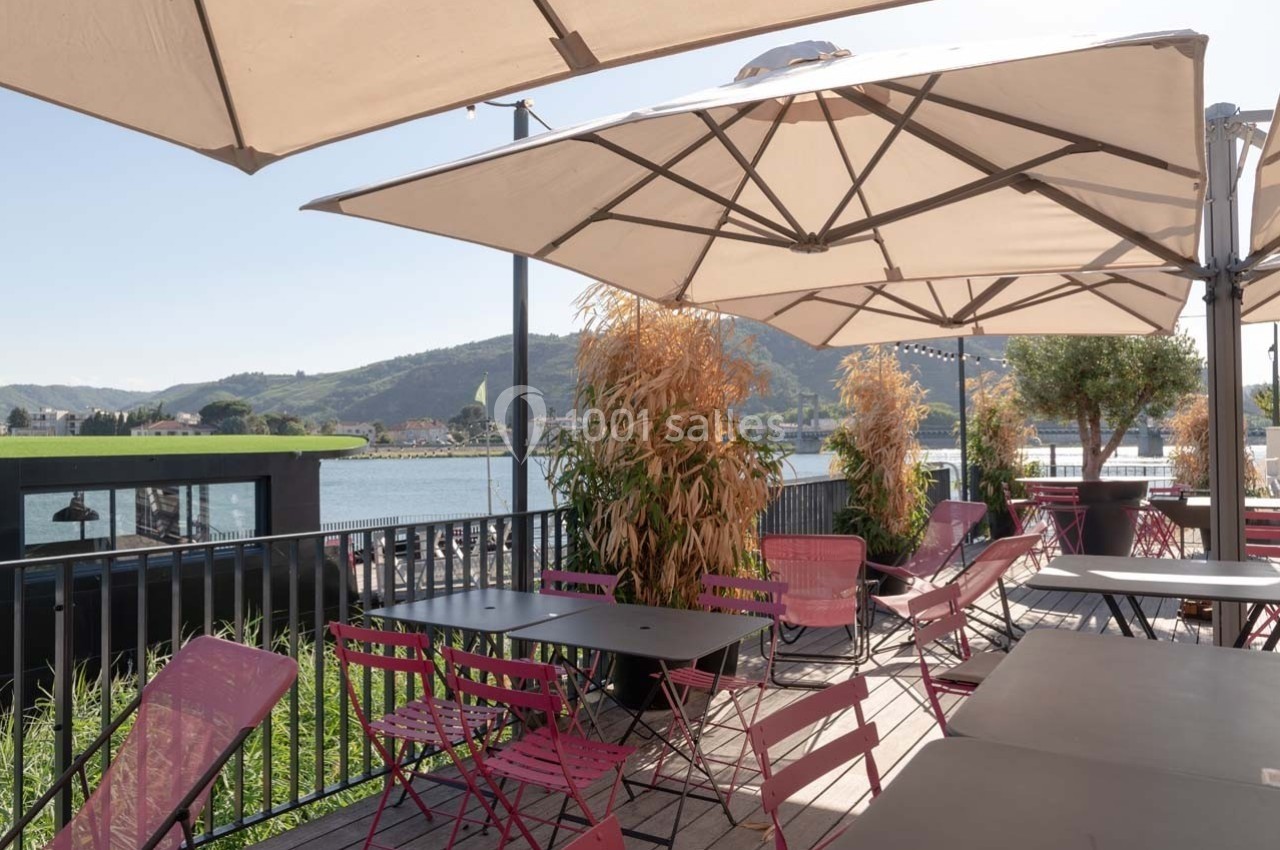 Terrasse ensoleillée avec tables et chaises colorées, vue sur un paysage de colline et un plan d'eau en arrière-plan.