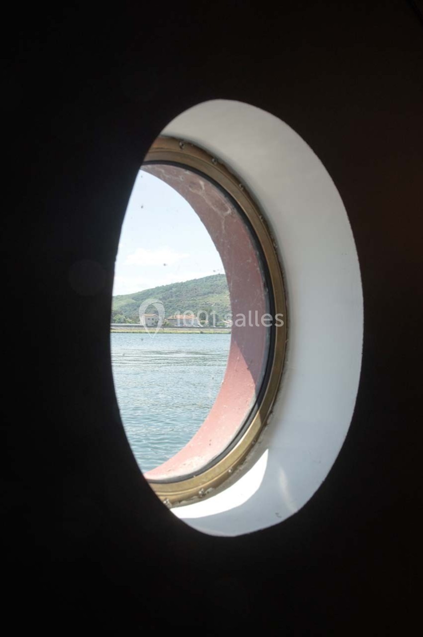 Vue d'une colline et d'un bâtiment au bord de l'eau à travers un hublot circulaire.