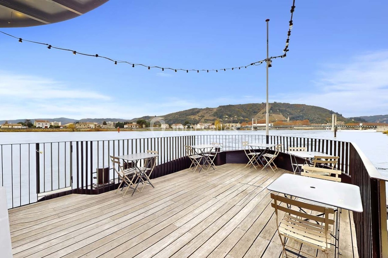 Terrasse en bois d'un bateau avec tables et chaises, vue sur une rivière, des collines et des habitations au loin.