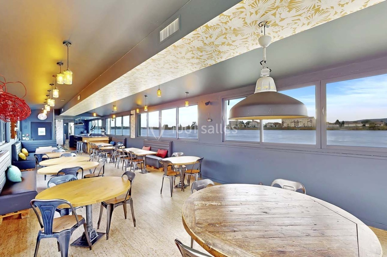 Salle lumineuse avec grandes baies vitrées donnant sur un paysage, tables en bois et chaises métalliques.