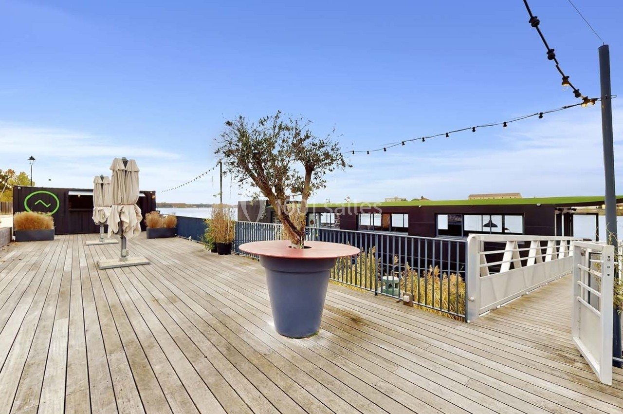 Terrasse en bois avec un grand pot d'arbre, des parasols fermés, des guirlandes lumineuses et vue sur un plan d'eau.