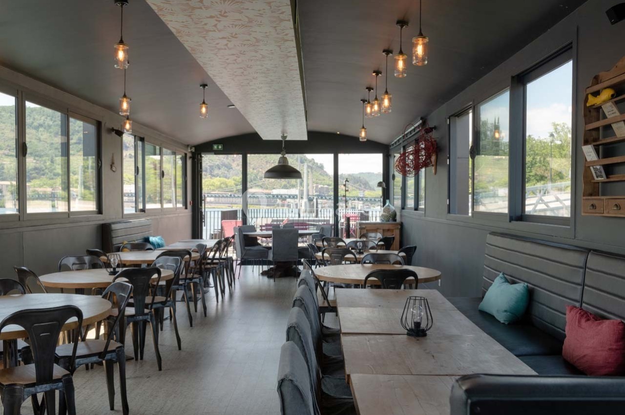 Salle de restaurant lumineuse avec grandes baies vitrées, tables en bois, chaises noires et vue sur un paysage extérieur.