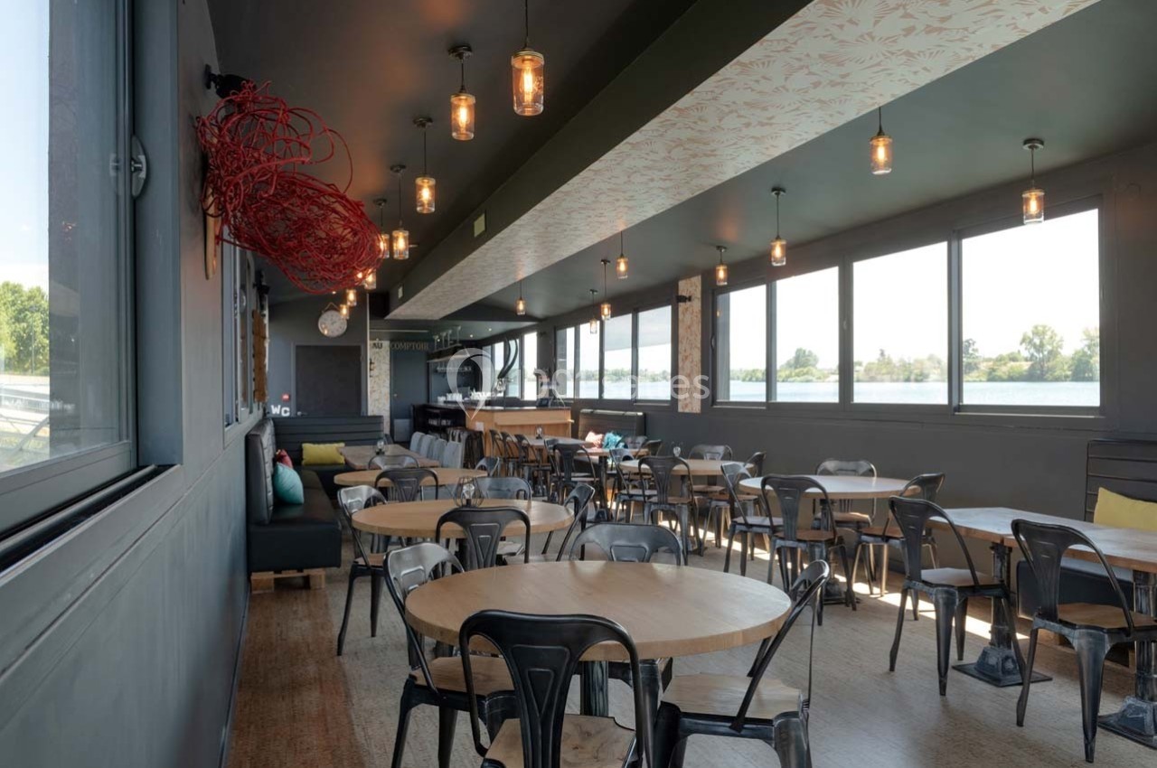 Salle de restaurant lumineuse avec des tables en bois, des chaises noires et de grandes fenêtres donnant sur un paysage…