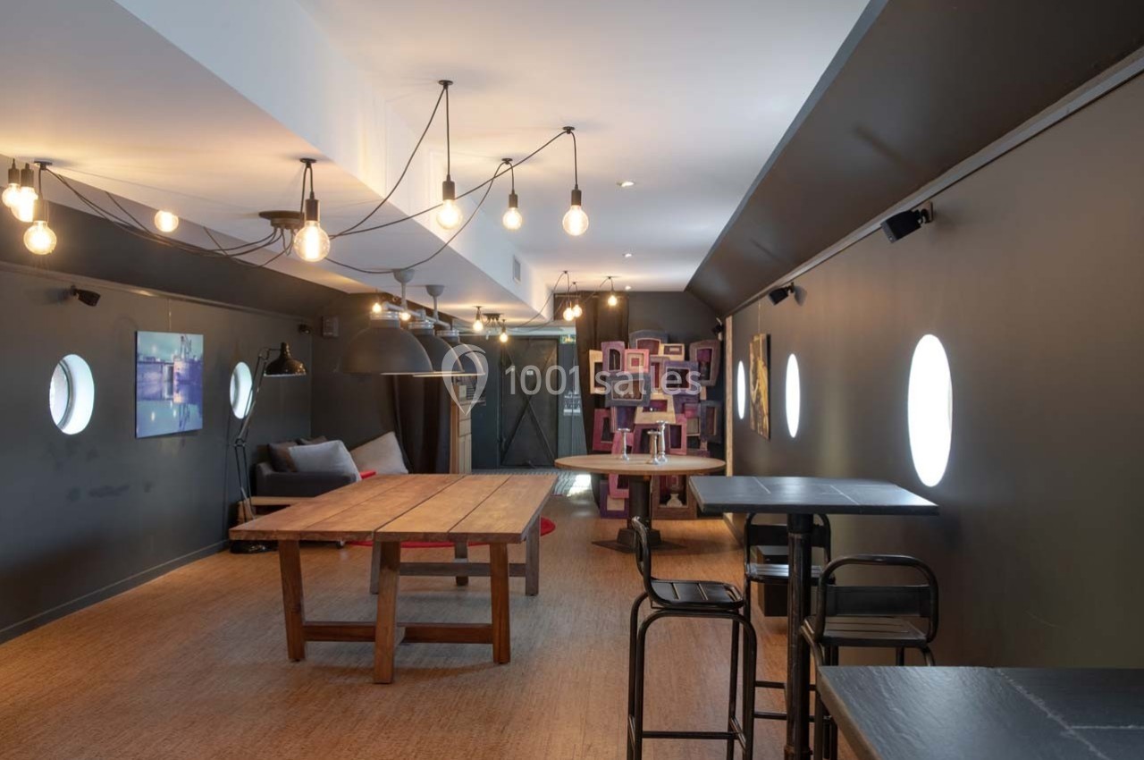 Salle moderne avec tables en bois, chaises noires, éclairage suspendu et décoration murale colorée au fond.