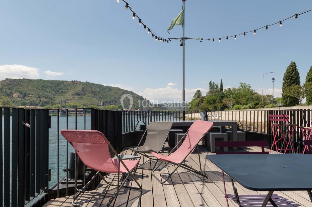 Terrasse en bois avec chaises longues et tables, vue sur une rivière et des collines sous un ciel dégagé.