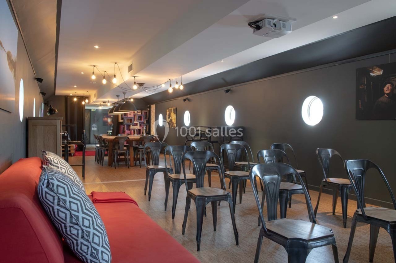 Salle lumineuse avec chaises en métal, canapé rouge au premier plan et décor moderne avec éclairage suspendu.