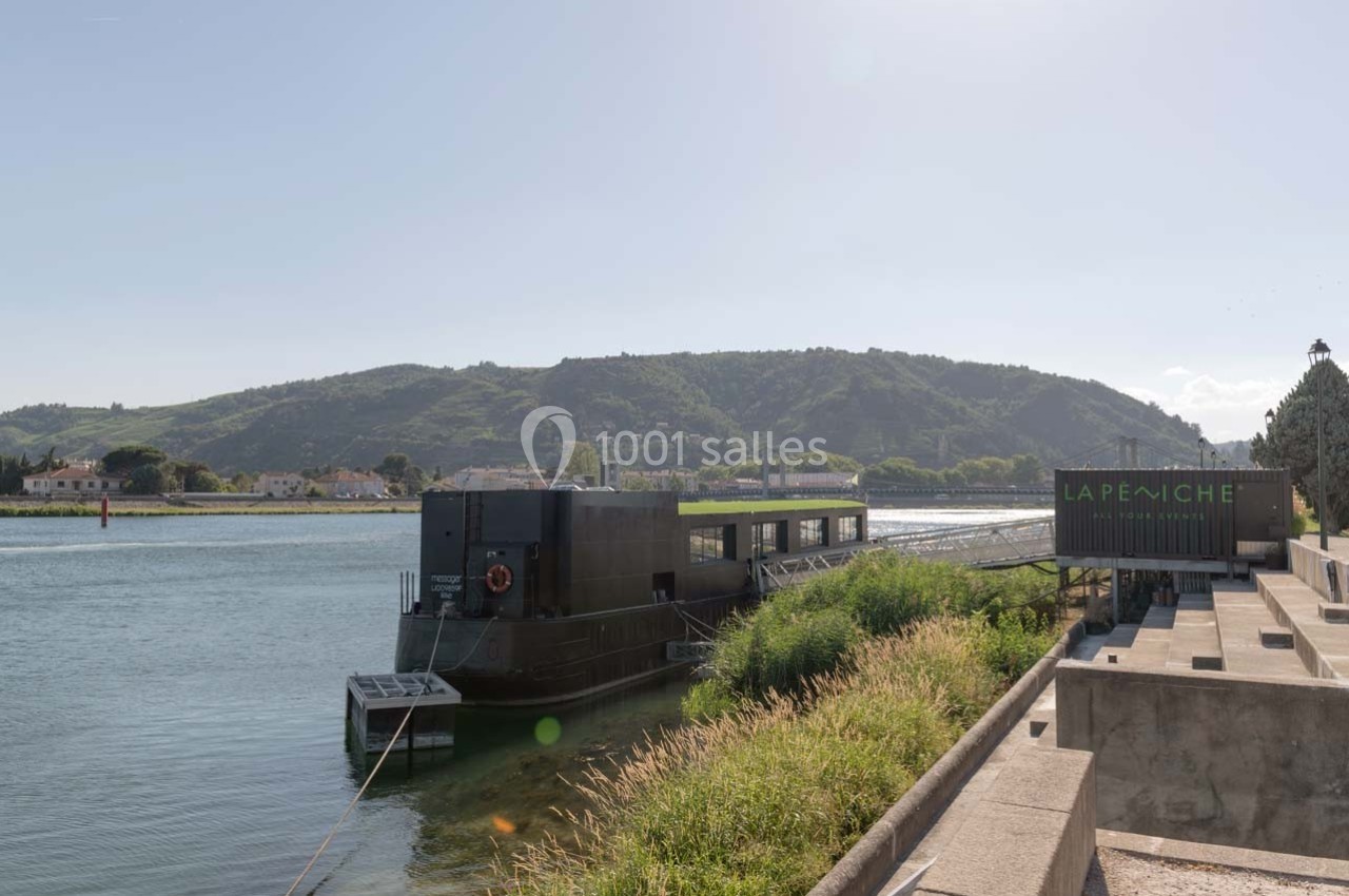 Bâtiment flottant noir amarré sur une rivière, entouré de verdure, avec des collines en arrière-plan.