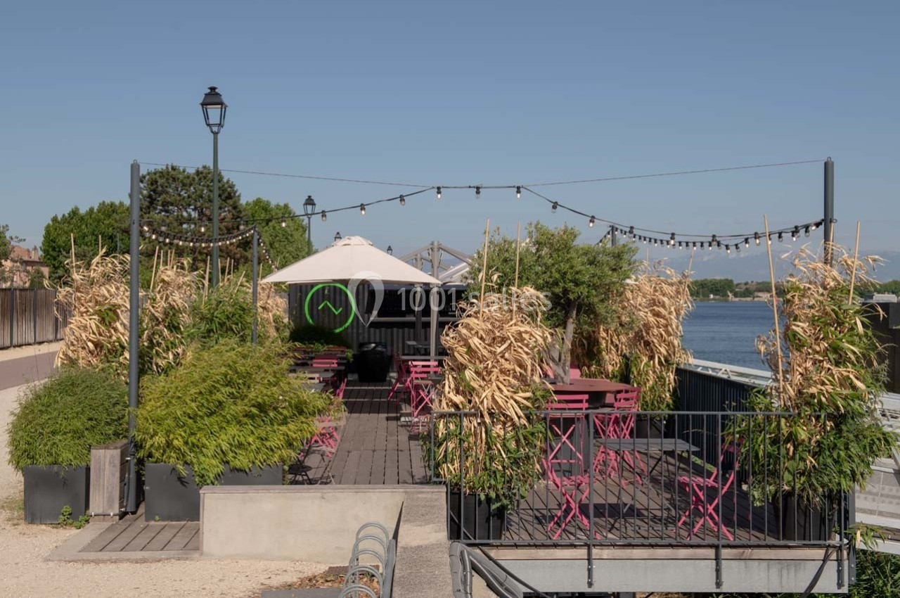 Terrasse extérieure avec tables et chaises roses, décorée de plantes et éclairée par des guirlandes, près d'un plan d'eau.