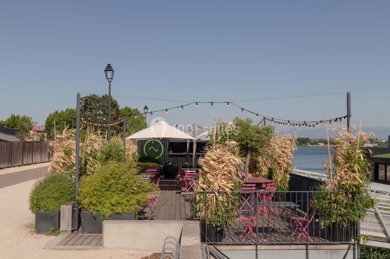 Terrasse extérieure avec tables et chaises roses, entourée de plantes, située près d'un plan d'eau sous un ciel dégagé.