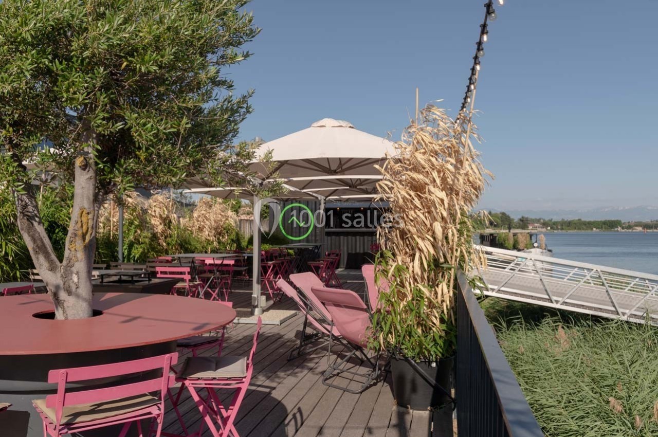 Terrasse ensoleillée avec tables et chaises roses, parasol blanc, végétation et vue sur un plan d'eau.