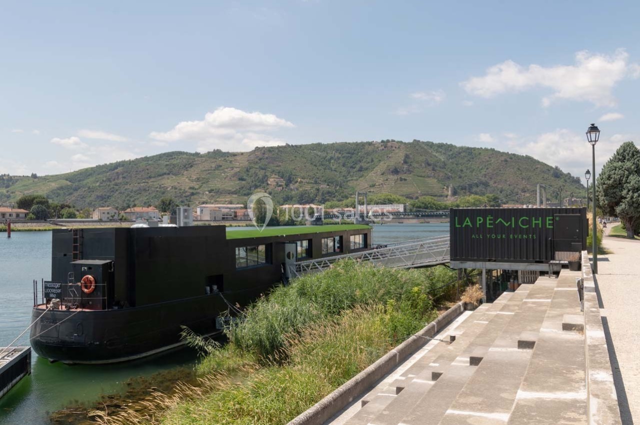 Bateau noir amarré sur un quai, entouré de verdure, avec des collines en arrière-plan sous un ciel partiellement nuageux.