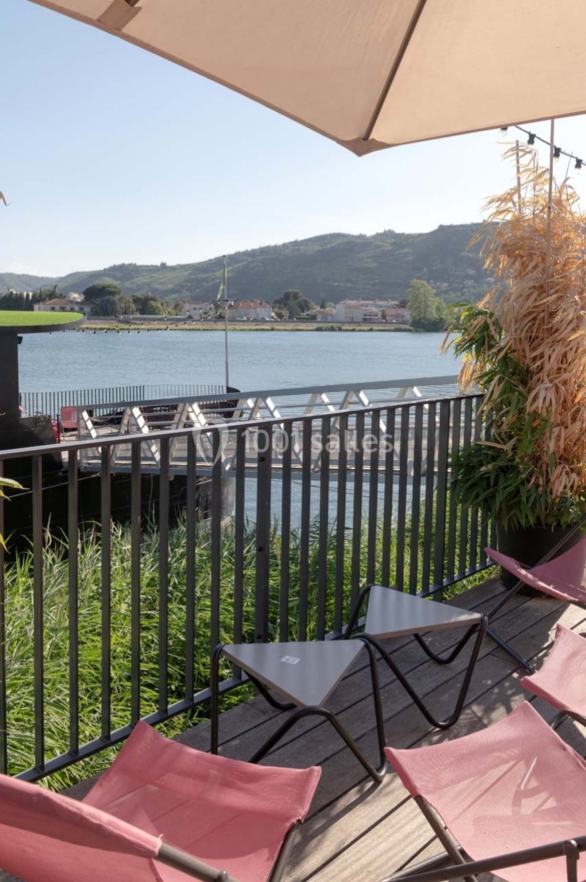 Terrasse avec chaises longues, vue sur un fleuve, des collines et un ponton, sous un grand parasol beige.