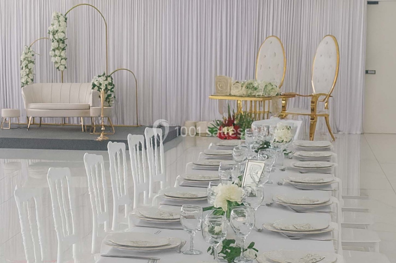 Salle de réception décorée pour un mariage avec table dressée, chaises blanches et espace pour les mariés en arrière-plan.
