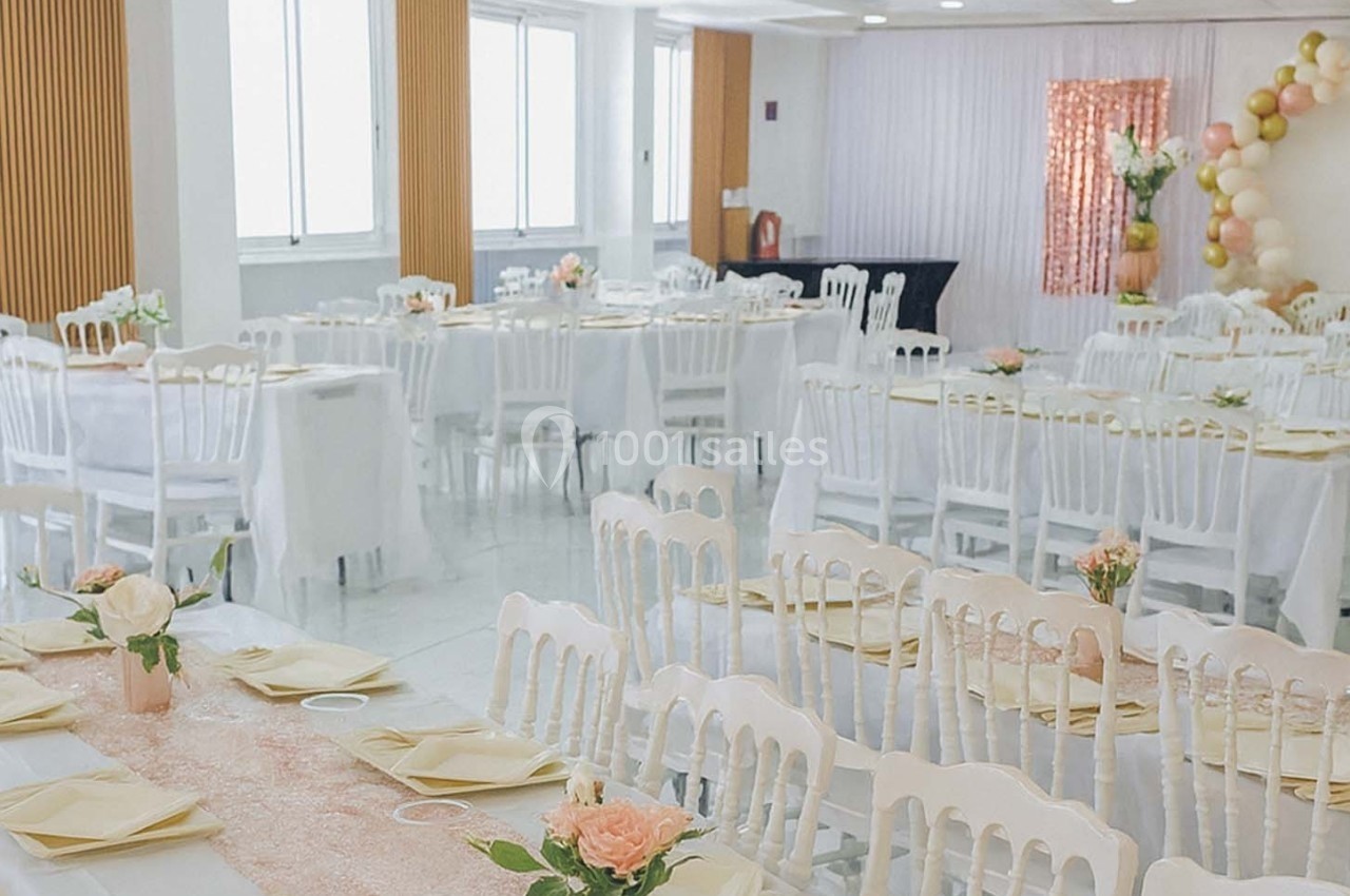 Salle de réception lumineuse décorée avec des nappes blanches, des fleurs et des ballons dans des tons pastel.