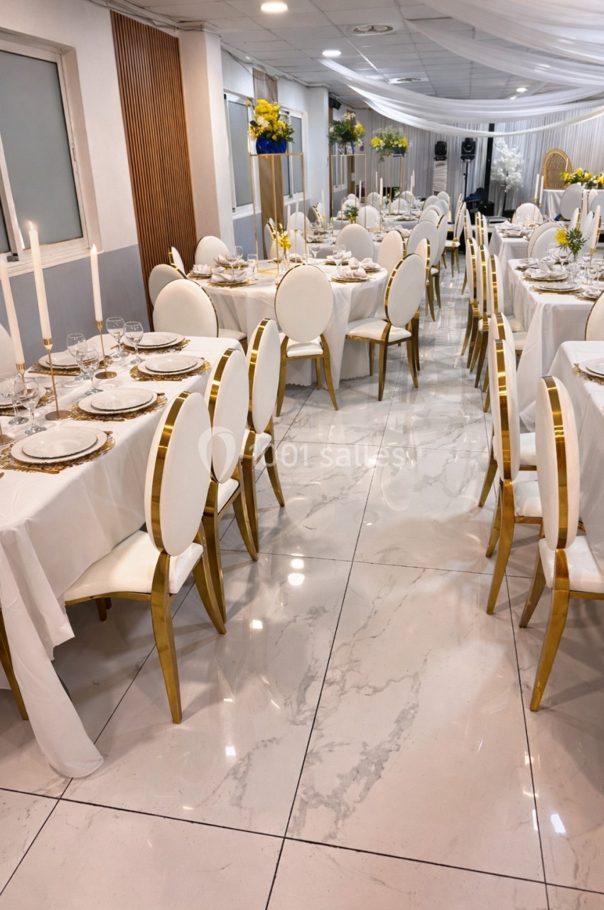 Salle de réception élégante avec tables dressées, chaises blanches et dorées, décorations florales jaunes et blanches.