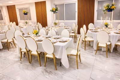Salle de réception avec tables dressées, nappes blanches, chaises dorées et arrangements floraux jaunes et blancs.
