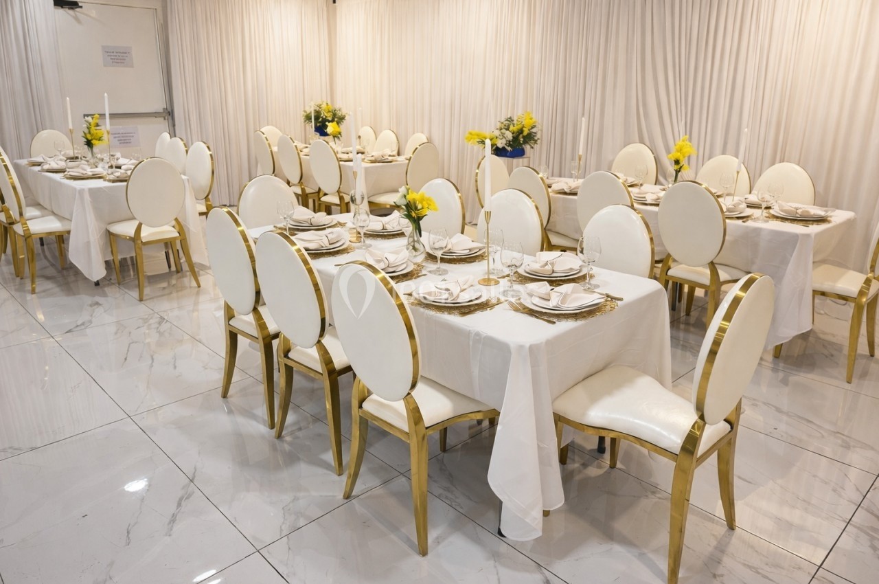 Salle de réception avec tables dressées, nappes blanches, chaises dorées et décorations florales jaunes.