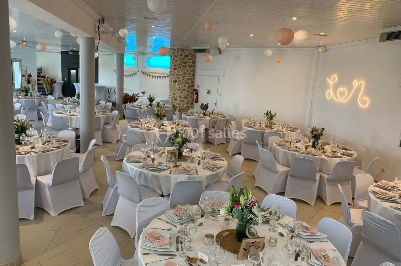 Salle de réception décorée pour un mariage, avec des tables rondes dressées, fleurs et guirlandes lumineuses.