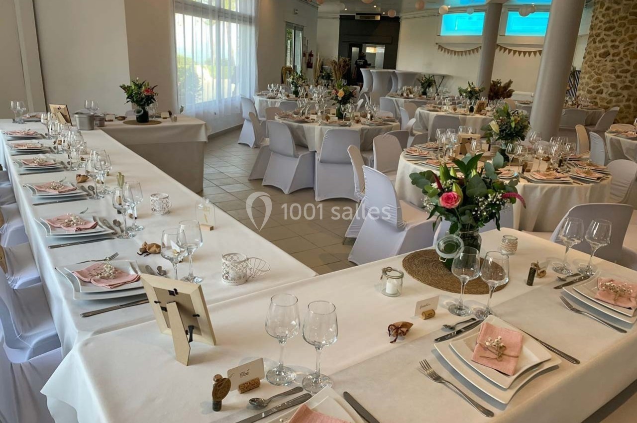 Salle de réception décorée pour un événement, avec tables dressées, nappes blanches et arrangements floraux.