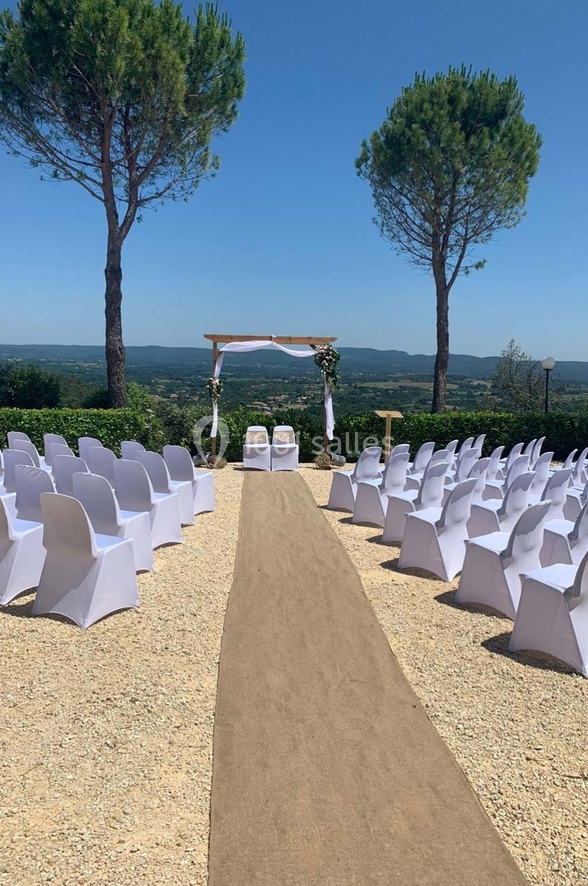 Allée centrale menant à une arche de mariage décorée, entourée de chaises blanches alignées en extérieur.