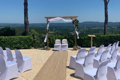 Chaises blanches alignées en extérieur face à une arche décorée, avec vue sur un paysage vallonné et des arbres.