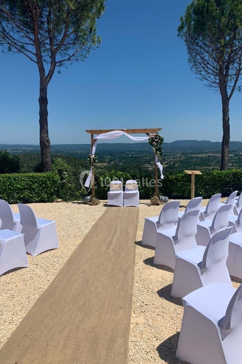 Allée centrale menant à une arche de mariage décorée, entourée de chaises blanches en extérieur avec vue panoramique.