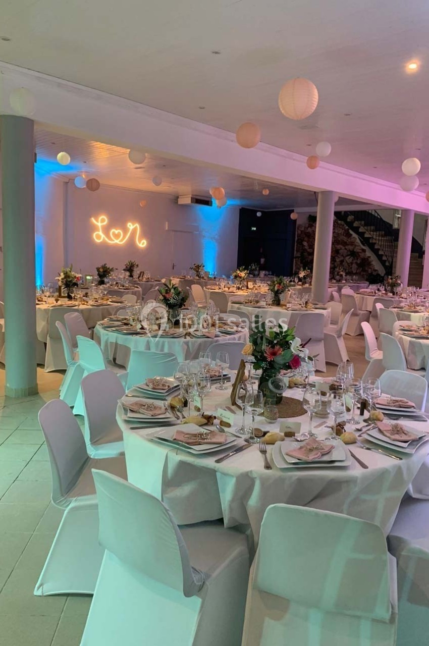 Salle de réception décorée avec des tables rondes dressées, des fleurs et des lanternes suspendues.
